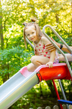 Young girl with light brown hair wearing a pink tank top and shorts sits on a red slide while an adult hand supports her from behind in a sunny park setting