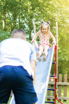 Young girl in pink dress with heart patterns slides down a playground slide while a boy in blue shorts watches from below in a sunny park setting
