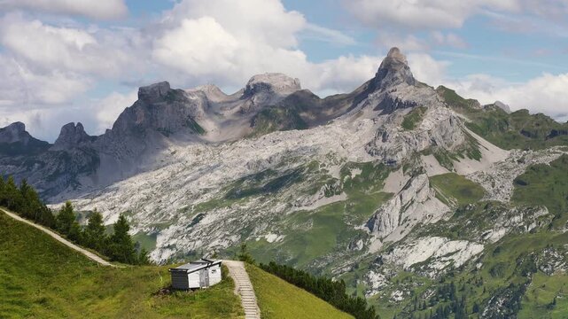 Rocky limestone peaks seen from the Klingenstock ridge near Stoos, Switzerland. Static shot of an alpine hut and hiking path on a green slope below grey summits in the Swiss Alps in summer