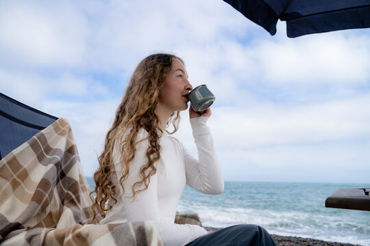 Woman relaxing with a hot beverage beside the sea during the cool season. Peaceful coastal lifestyle, wellness and mindful leisure concept.
