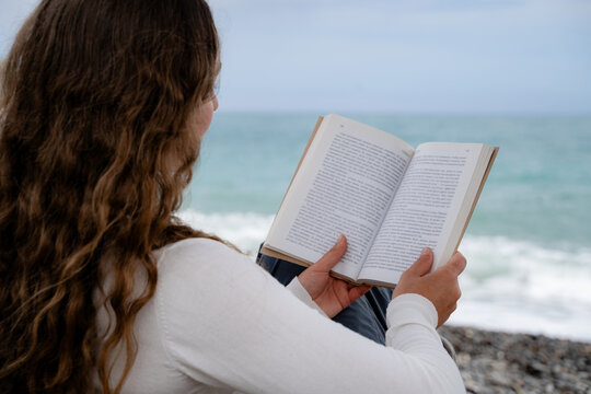A woman is enjoying a printed book while sitting by the ocean, back view. The calm seaside atmosphere reflects the digital detox in nature and the slow living concept.
