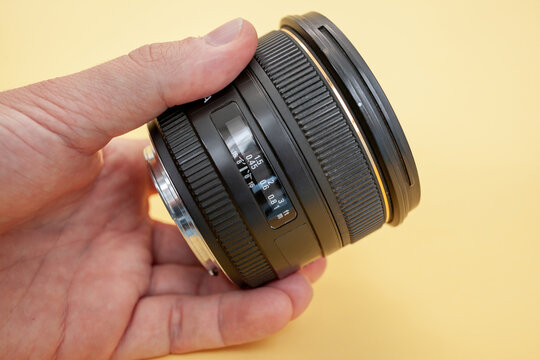 Camera lens is being held in a hand, showcasing the focus ring and aperture markings, set against a bright yellow background