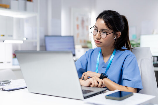 Healthcare professional in blue scrubs working on laptop in medical office, representing digital, patient records, clinical work, telehealth, nursing and medical technology.