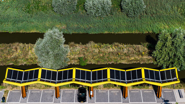 Aerial view of a modern solar-powered parking structure with a zigzag roof, wooden columns, and a car parked beside a canal with lush vegetation in Den Hoorn, South Holland, Netherlands.