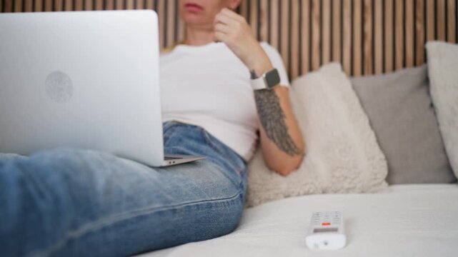 Woman working on laptop from bed at home before using an air conditioner remote control to cool the room, illustrating hot summer weather, remote work and indoor comfort