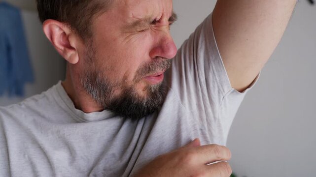 Bearded man with a disgusted expression smelling his sweaty t-shirt due to unpleasant underarm odor, illustrating excessive perspiration, body odor and personal hygiene