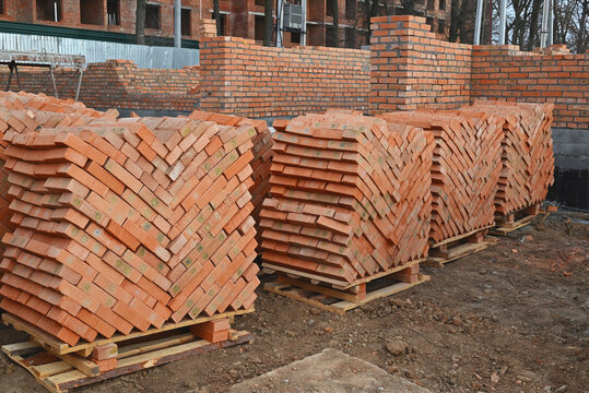 Large stacks of red clay bricks on wooden pallets at a construction site. An unfinished building is visible in the background.