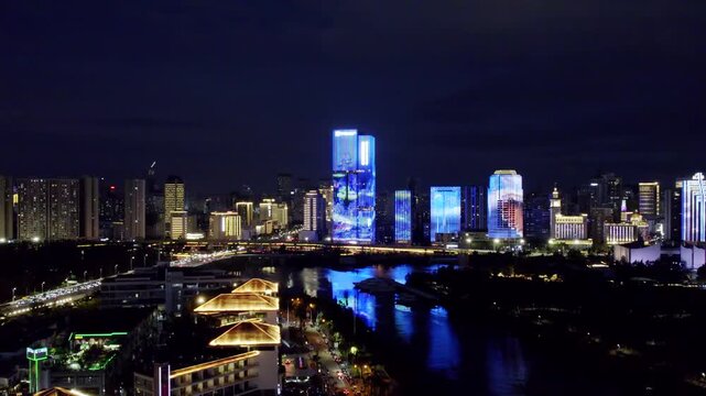 A night urban landscape featuring illuminated modern buildings, a river with light reflections, and a busy road along the waterfront.