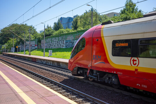 Warsaw, Masovian, Poland - July 30, 2026: A modern red and cream SKM passenger train moving along the railway track past an open platform.