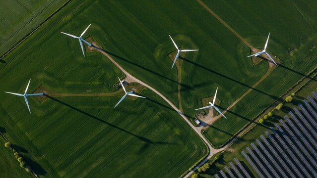 Top down aerial view of wind turbines casting long shadows across green agricultural fields with adjacent solar panel farm, sustainable renewable energy infrastructure generating clean energy from ab