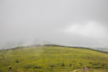 Fototapeta premium Lone hiker stands on a vast green grassy hill under a moody foggy mountain landscape at dawn