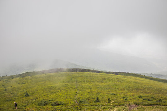 Lone hiker stands on a vast green grassy hill under a moody foggy mountain landscape at dawn