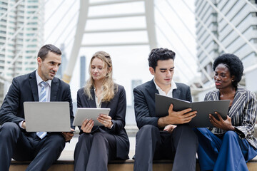 Obraz premium Diverse business team in formal attire working outdoors on laptops and tablets, collaborating on a project in a modern urban setting with architectural backdrop.