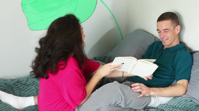 Young man and woman spending a relaxing evening together while reading a book in bed. Their natural conversation and affectionate interaction represent romance, happiness. Mid shot, top view.