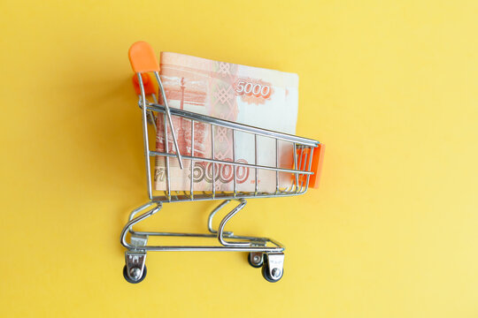 Mini shopping cart contains 5000 ruble banknotes against a vibrant yellow background, illustrating concepts of shopping and finance