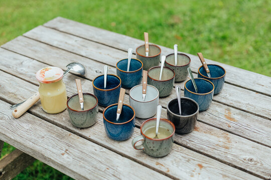 Assorted ceramic cups with spoons arranged on a rustic wooden table beside a jar of pale herbal drink, set outdoors on green grass