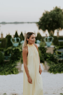 Woman wearing yellow dress stands on beach at sunset near gardens and water