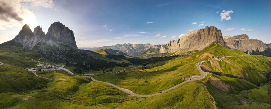 Aerial view of the winding road at Sella Pass surrounded by lush green mountain slopes and the dramatic rocky peaks of the Dolomites in Selva, Trentino-Alto Adige, Italy.
