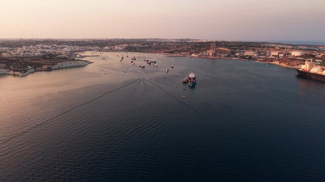 Marsaxlokk Port, Malta, at golden hour, with San Lucjan Tower, oil tanker, fuel dolphin, LNG terminal and boats crossing the Mediterranean Sea in the sunset.