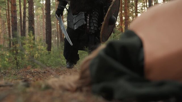 Extreme low-angle motion shot showing armored figure stepping forward past prone person lying on pine-needle covered ground; visible action is confident stride of warrior holding raised sword and