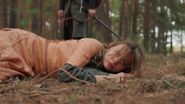 Young woman in period dress lies on pine needle covered forest floor and pushes up on one elbow while standing figure nearby holds rope and watches, suggesting tense woodland rescue or confrontation