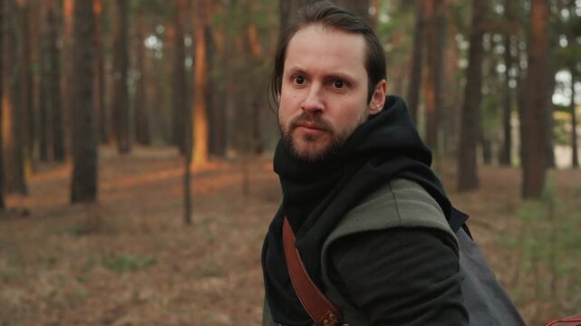 Bearded man in layered dark clothing turns upper body in pine forest, looking overshoulder with focused expression while holding bow low across chest, showing alert scouting movement