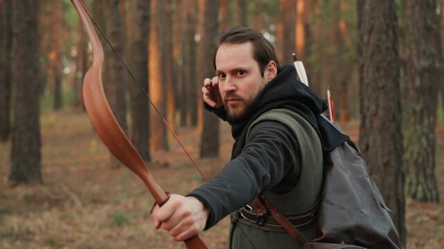 Middle-aged man in medieval archer costume draws wooden longbow, holds taut bowstring, releases shot, then lowers bow and pauses among pine trees with focused contemplative expression