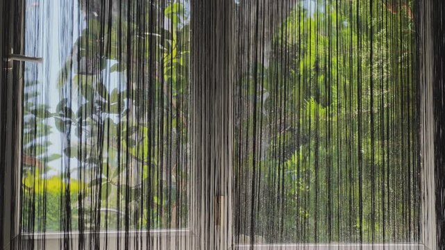 Thread curtain moving in the wind with a bright, sunny garden outside the window. Peaceful and relaxing interior view on a beautiful day