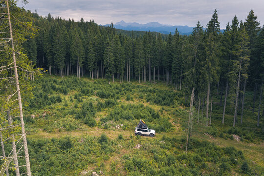 Aerial view of a white SUV with a roof tent parked in a clearing surrounded by a dense pine forest with distant mountains in the background in Slovenia.