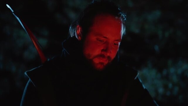 Cloaked man with longbow lowers head and looks downward under red and blue night lighting, showing weary focus, quiet tension, dark costume detail, and cinematic character mood, with man, archer