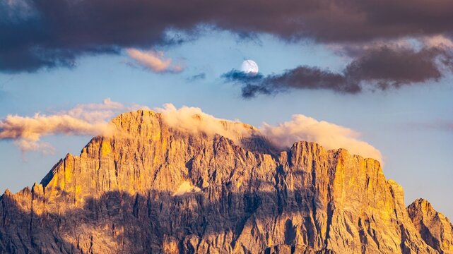 Aerial view of Monte Civetta illuminated by golden hour light with clouds and a visible moon in the sky in Alleghe, Veneto, Italy.
