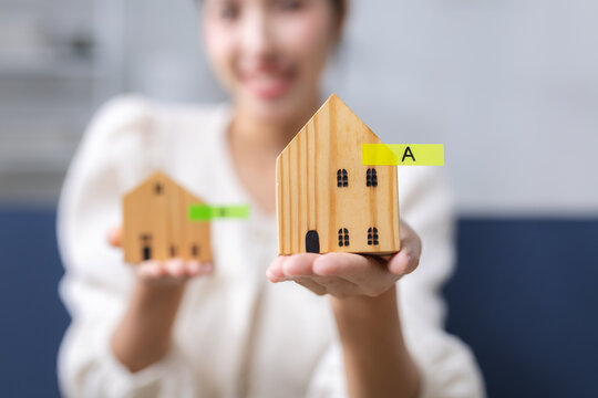 Asian woman holding two wooden house models for comparison, representing home buying, real estate choice, property selection, housing investment, mortgage planning and ownership decision.