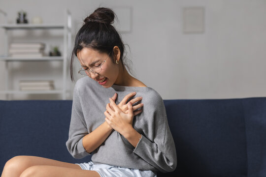 Woman holding chest in pain while sitting on sofa at home, showing sudden discomfort, heart health concern, chest pressure, illness symptoms, medical emergency, healthcare and wellness concept.