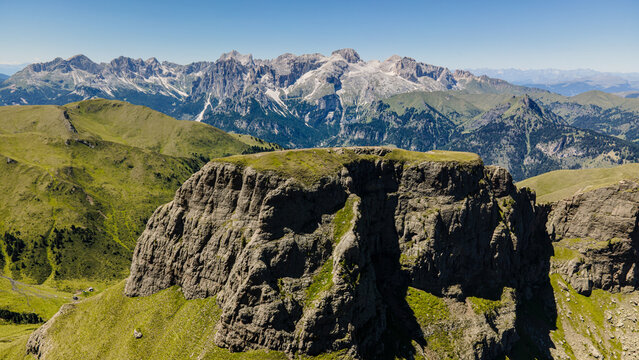Aerial view of the rocky Crepa Neigra peak surrounded by lush green alpine slopes and the jagged Dolomites mountain range under a clear blue sky in Canazei, Trentino-Alto Adige, Italy.