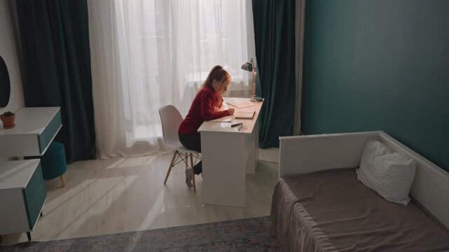 Young woman in red top is seated at white desk writing. Room features teal walls dark green curtains partially drawn. Bed with gray bedding sits nearby and various study materials