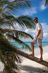 Wearing sunglasses, a man stands on a curved palm tree trunk along a sandy tropical beach in Maldives by turquoise ocean water under a sunny sky. © Aerial Film Studio