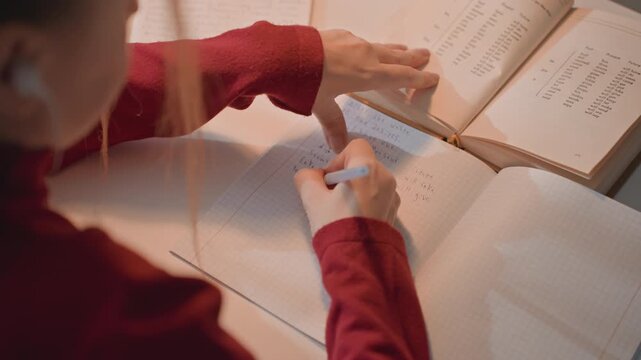 Young woman in red top writes with blue pen on graph paper referencing. Her hands and notebook are captured closely. Scene is set at desk under warm indoor lighting focusing solely