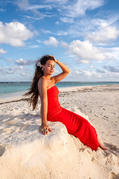 A woman wearing a strapless gown sits on the white sand bank along the shoreline of Dhigurah island in Maldives under a partly cloudy sky.
