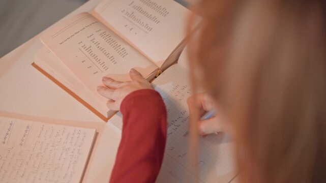 Young woman with light brown hair studies grammar. Woman wears red sleeve and interacts with study materials. Scene is focused on close up of hands notebooks pen book text