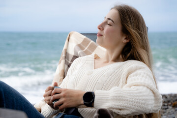 A young woman with closed eyes, wearing a casual sweater, sits by the sea, enjoying the fresh ocean air on a cool, cloudy day, experiencing mental restoration and peace of mind in nature.