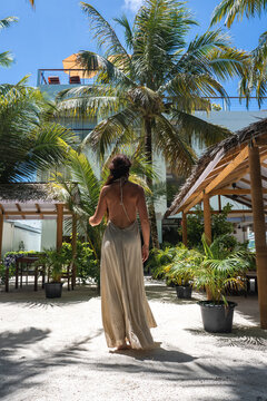 A woman wearing a backless long dress walks along a sandy path lined with palm trees and gazebos at a tropical hotel resort in Dhigurah island, Maldives.