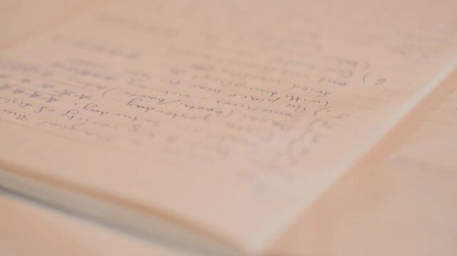 Close up of open notebook with handwritten notes. Camera moves steadily rightward over cream colored lined paper filled. Soft indoor lighting illuminates neutral toned desk surface