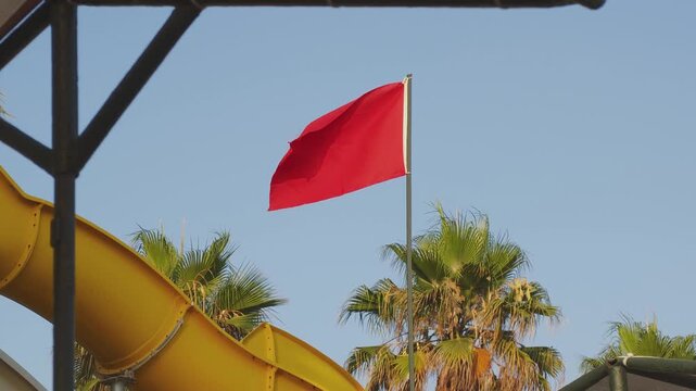 Red flag fluttering in the wind against a clear blue sky. A yellow water slide and green palm trees are visible in a sunny water park