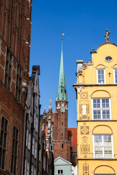 The tall spire of Saint Mary church rises between decorated historic townhouse facades in the old town of Gdansk, Poland under a clear sky.