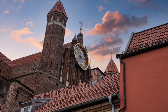 Brick gothic tower and dial of Saint Mary church rise above tiled rooftops under sunset clouds in the historic district of Gdansk, Poland.