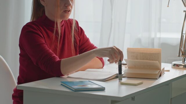 Young woman in red top sits at white desk interacting. Woman manipulates open book propped upright. Scene is set in bright room with sheer curtains featuring various study