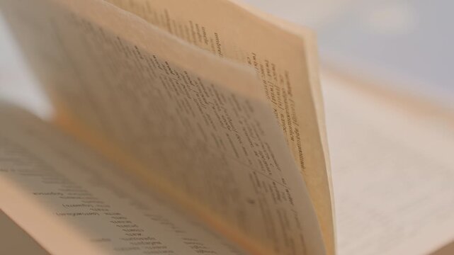 Close up of open book with text camera moves downward. Pages turn from bottom left to top right as focus remains. Soft lighting creates warm tones against blurred background