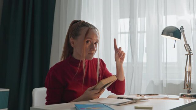 Young woman in red top reads book at white desk. Woman raises finger for emphasis then places closed brown book. Scene features sheer curtains dark green drapes metallic lamp