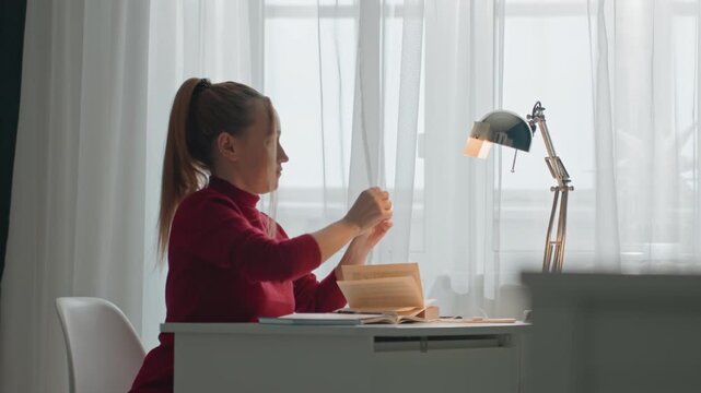Young woman in red turtleneck sits at white desk interacting. Woman moves her hands over open materials on cluttered workspace. Scene features sheer curtains bright natural light