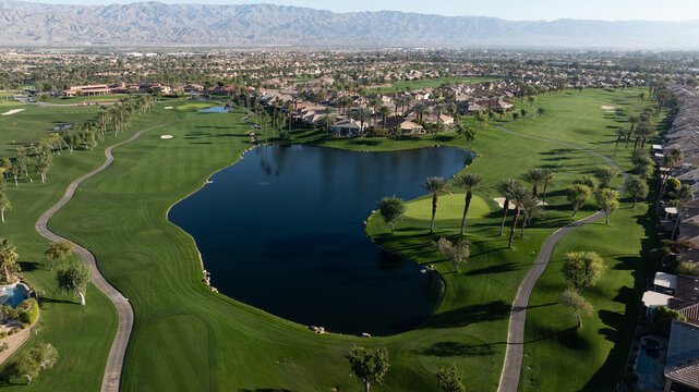 Aerial view of a golf course with a large central pond, manicured fairways, palm trees, and surrounding residential homes with mountains in the distance in Indio, California, United States.